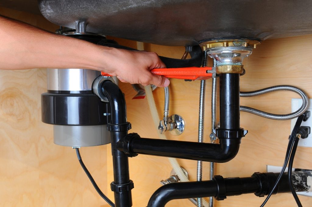 Closeup of a plumber using a wrench to tighten a fitting beneath a kitchen sink. Only the man's hand and arm are visible. Horizontal format.